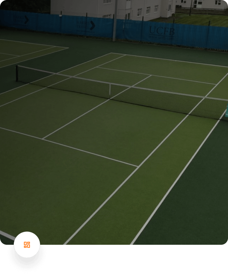 Empty tennis court with green surface, surrounded by blue fence and buildings in the background.