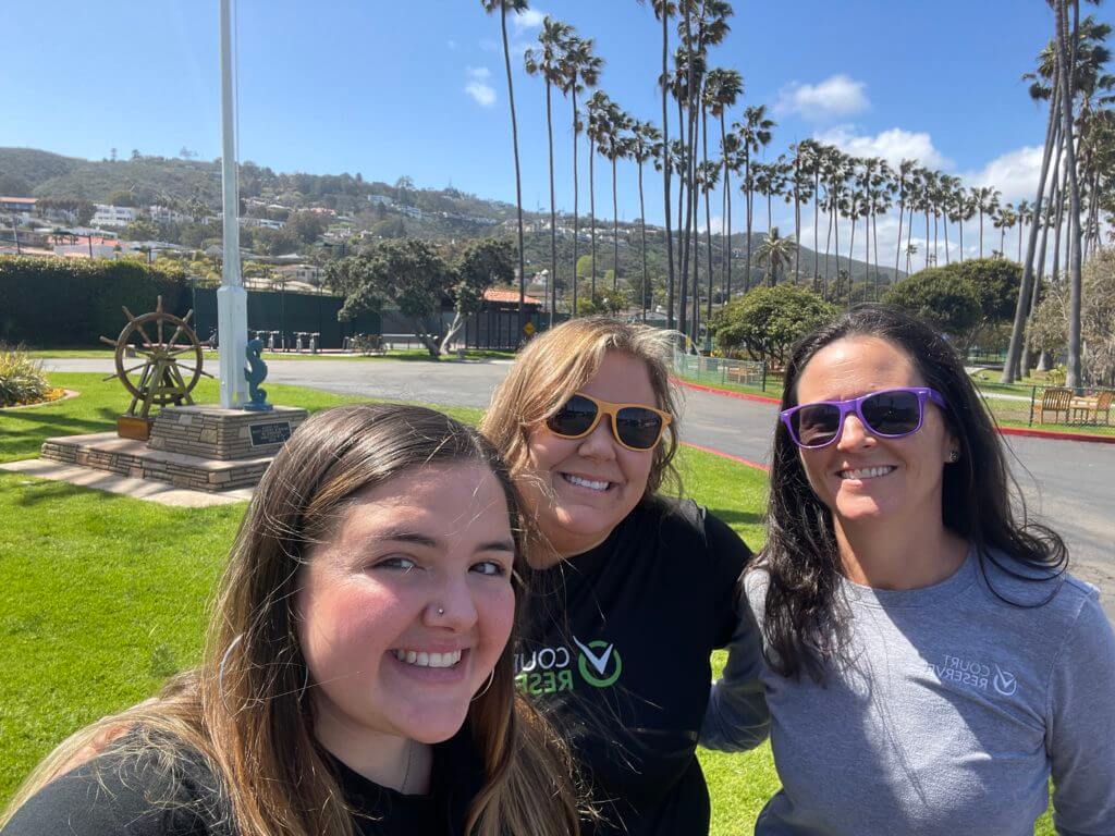 Three people smiling in a sunny park with palm trees and hills in the background.