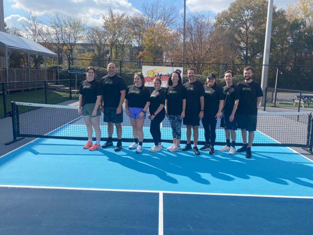 Nine people in matching black shirts on a blue pickleball court, trees and building in the background.