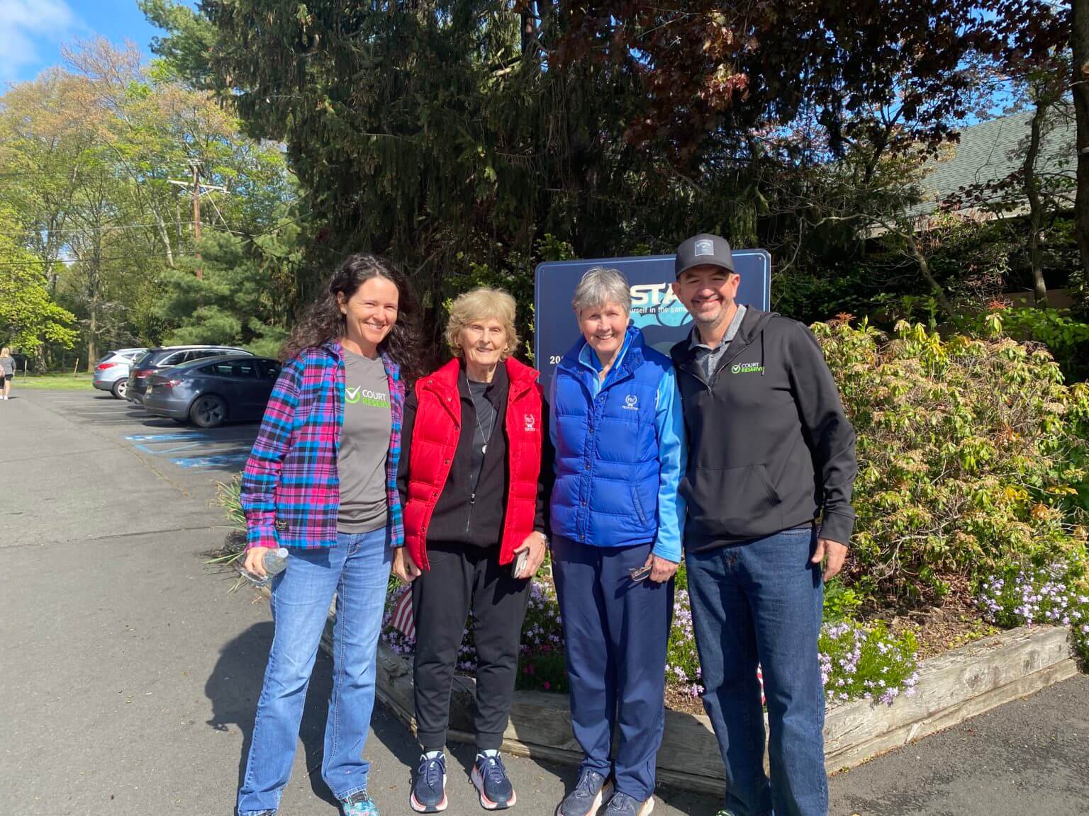 Four people smiling outdoors in front of a sign, with cars and trees in the background.