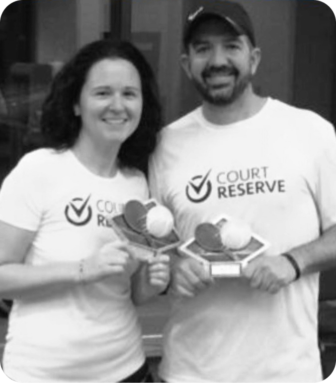 Two people in "Court Reserve" shirts smiling with trophies topped by paddles and a ball.