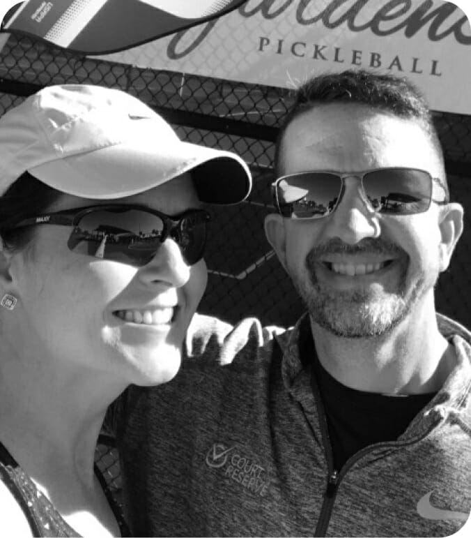 A smiling man and woman wearing sunglasses and casual sportswear in front of a pickleball sign.