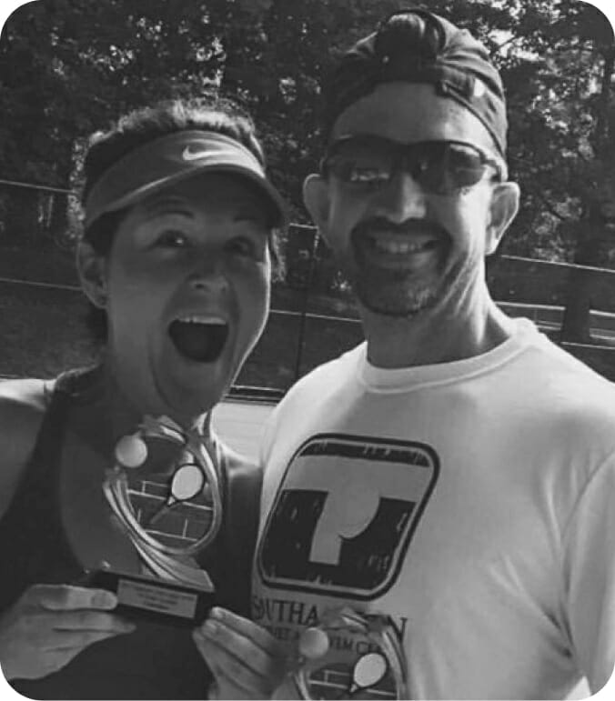 Two people smiling, holding tennis-themed trophies outdoors.