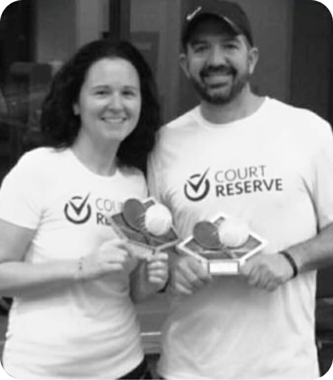 Two people smiling and holding trophies, wearing "Court Reserve" shirts.