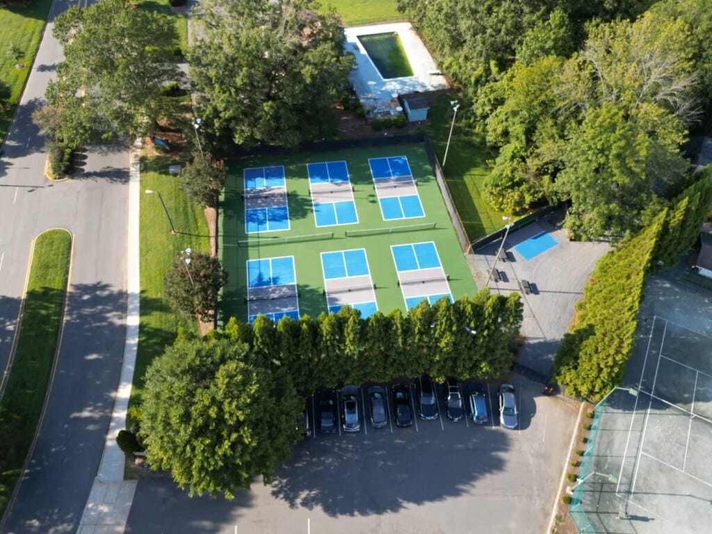 Aerial view of six blue-green tennis courts with adjacent parking lot and nearby buildings