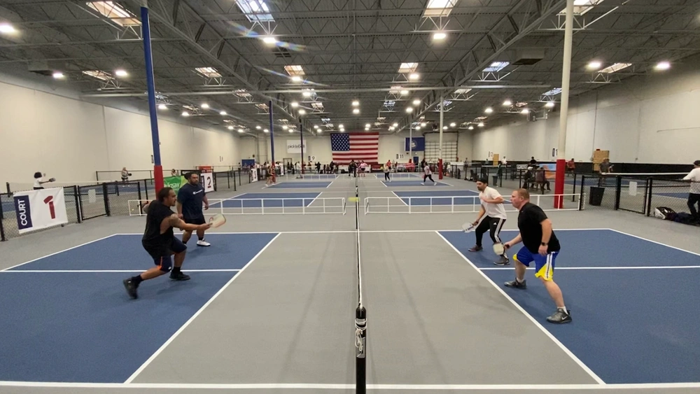 People playing pickleball on indoor courts with a large American flag hanging in the background.