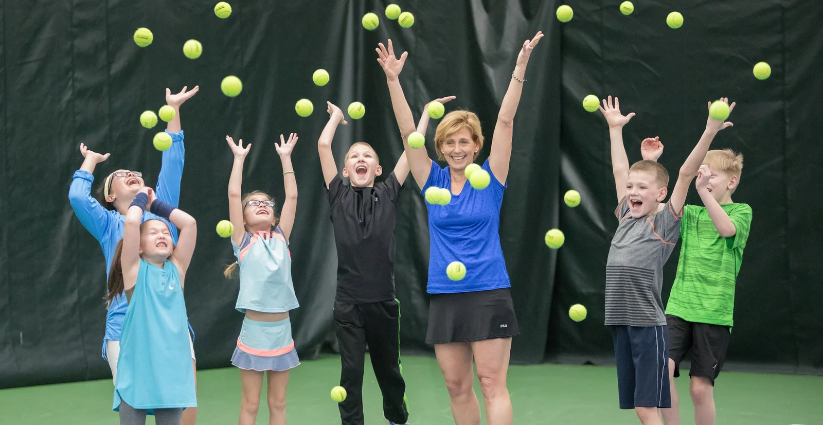 A group of children and an adult woman joyfully throwing tennis balls in the air indoors.