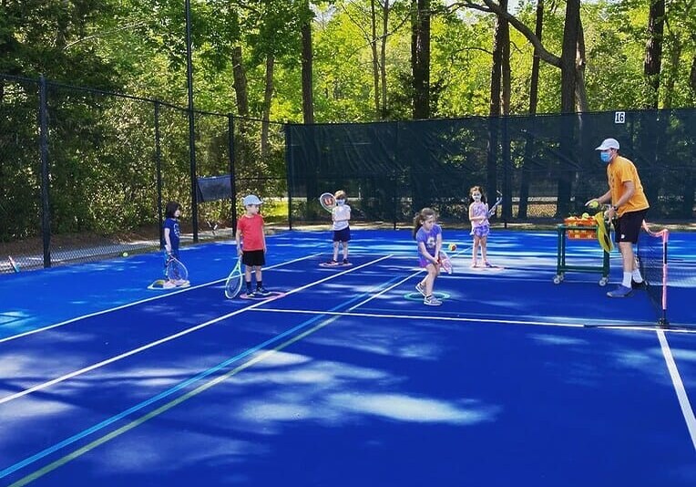 Children and a coach on a blue tennis court surrounded by trees during a tennis lesson