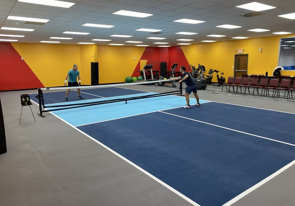 Two people playing pickleball indoors on a blue court with gym equipment in the background.