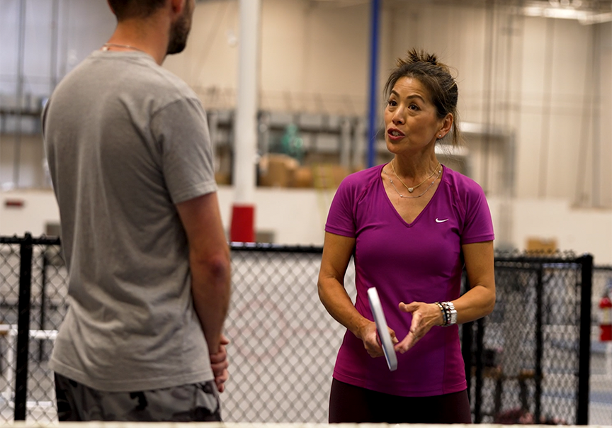 A woman in a purple shirt holds a tablet and talks to a man in a gray shirt in an indoor setting.