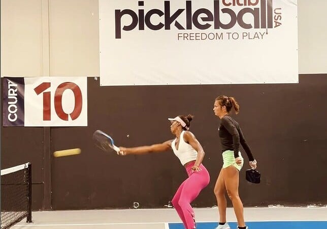 Two women playing pickleball in an indoor court, one swinging at the ball.